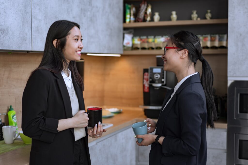 Two women in black and white formal suits chatting by a coffee vending machine in an office