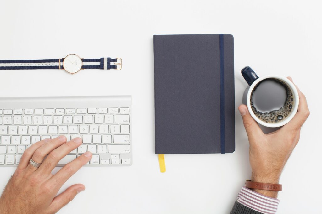 A man nursing a cup of coffee machine rental service coffee at a desk with a watch, a keyboard, and a blue notebook on it