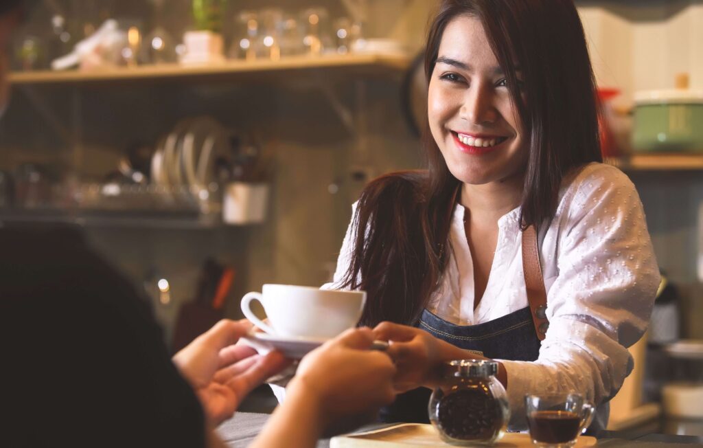 Barista wearing black and serving a smiling customer with a cup of coffee in a white coffee cup